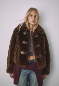 Hnedá bunda z umelého kožušiny s veľkými gombíkmi, so širokým golierom a skráteným strihom. Nosená cez pruhované tričko a modré rifle.