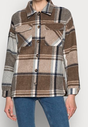 Femme portant une veste-chemise en laine à carreaux marron, beige et gris, boutonnée, avec deux poches poitrine, et un jean bleu, debout devant un fond uni.