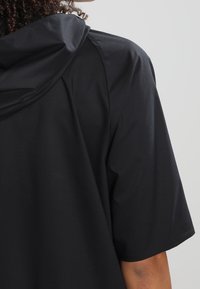 Černé tričko s krátkým rukávem a kapucí vyrobené z texturované látky, s volnějším střihem a jemným geometrickým vzorem na povrchu.