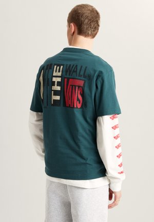 T-shirt a manica corta color verde acqua con grande logo "VANS" multicolore sul retro; indossata sopra una maglia bianca a manica lunga con testo rosso ripetuto.