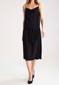 Schwarzes ärmelloses Kleid mit drapierter Halslinie, tailliert mit einem Gürtel, und einem fließenden Rock. Hergestellt aus einem glatten, leichten Stoff.