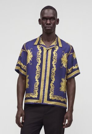 Homme portant une chemise bleue à manches courtes avec des motifs décoratifs dorés, debout devant un fond clair uni.