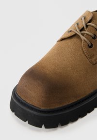 Ruskea mokkanahkakengät, joissa on pyöreä kärki, teksturoitu pinta ja musta kumipohja, jossa on sahalaitareuna. Varustettu ruskeilla nauhoilla ja nyöreillä.