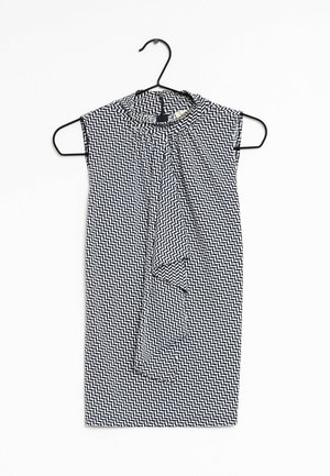 Robe sans manches à motif chevron noir et blanc avec un lien autour du cou, suspendue sur un cintre noir contre un fond blanc.