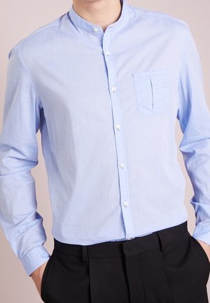 Man wearing a light blue mandarin collar shirt with buttons and a chest pocket, paired with black trousers, hands in pockets.