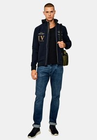 Felpa con zip blu navy con testo dorato, t-shirt nera, jeans blu e sneakers nere. Il modello porta una borsa verde, mostrando un look casual a strati.