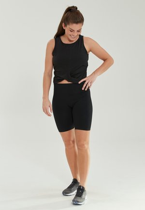 Svart träningsset bestående av en figursydd linne med vriden nederkant och högmidjade cykelshorts. Modellen bär svarta träningssneakers.