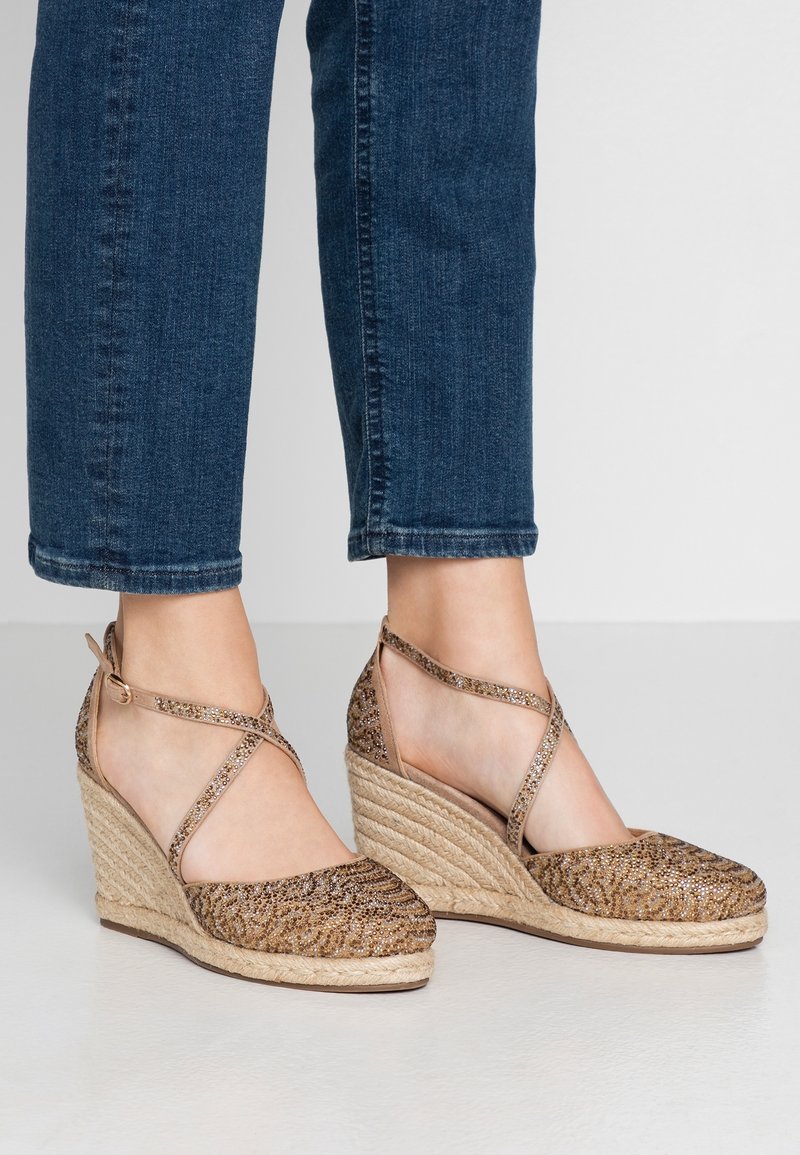 Tacones en cuña de color marrón claro con un patrón decorativo de purpurina, suela de yute trenzada y correas en el tobillo. Combinados con unos vaqueros oscuros.