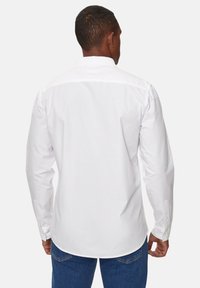 Camicia bianca a maniche lunghe con una texture liscia, colletto con bottoni e dettaglio sul polsino. Orlo leggermente curvo, design semplice e classico.