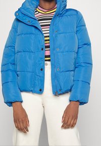 Ljusblå puffjacka med huva, med dragkedja och knappstängning, sidofickor och kviltad textur. Bärs över en randig topp.