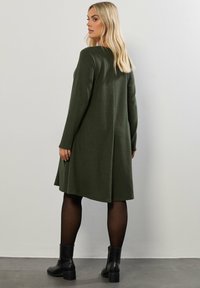 Knee-length, olivgrön tröjklänning med långa ärmar, en avslappnad passform och textilstruktur. Buren med svarta ankelstövlar och strumpbyxor.