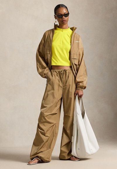 Καφέ ελαφρύ μπουφάν με φερμουάρ, συνδυασμένο με φωτεινό κίτρινο κομμένο τοπ και καφέ παντελόνια που ταιριάζουν. Άσπρη τσάντα τύπου slouch και μαύρα γυαλιά ηλίου.