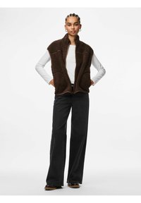 Rudas pūkų liemenė su aukštu apykakle, užtrauktuku ir dviem priekinėmis kišenėmis. Dėvima ant balto ilgomis rankovėmis marškinio ir derinama su juodais plačiais kelnėmis.