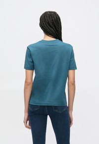 Tumesinise-roheline lühikeste varrukatega T-särk ümmarguse kaeluse, pehme tekstuuri ja sirge lõikega, kantud tumeliste teksadega.