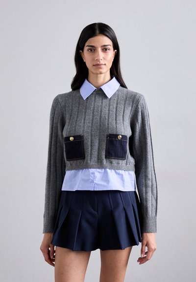 Gray ribbed sweater with two navy pocket accents featuring gold buttons, layered over a light blue collared shirt and paired with a navy pleated skirt.