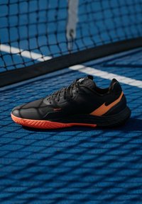 Μαύρο αθλητικό παπούτσι με πορτοκαλί λεπτομέρειες, υφή στην επάνω επιφάνεια, μαξιλαρωτή σόλα και σχέδιο ζιγκ-ζαγκ στην εξωτερική σόλα, σε μπλε φόντο.