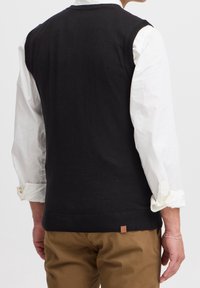 Gilet in maglia nera con finiture a coste, presenta un collo rotondo ed è indossato sopra una camicia bianca a maniche lunghe, abbinato a pantaloni marroni.
