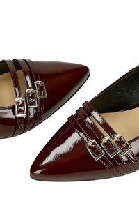 Glanzende donkerbruine platte schoenen met een spitse neus, drie dunne bandjes met zilveren gespen over de bovenzijde, een beige voering en zwarte rand langs de zolen.