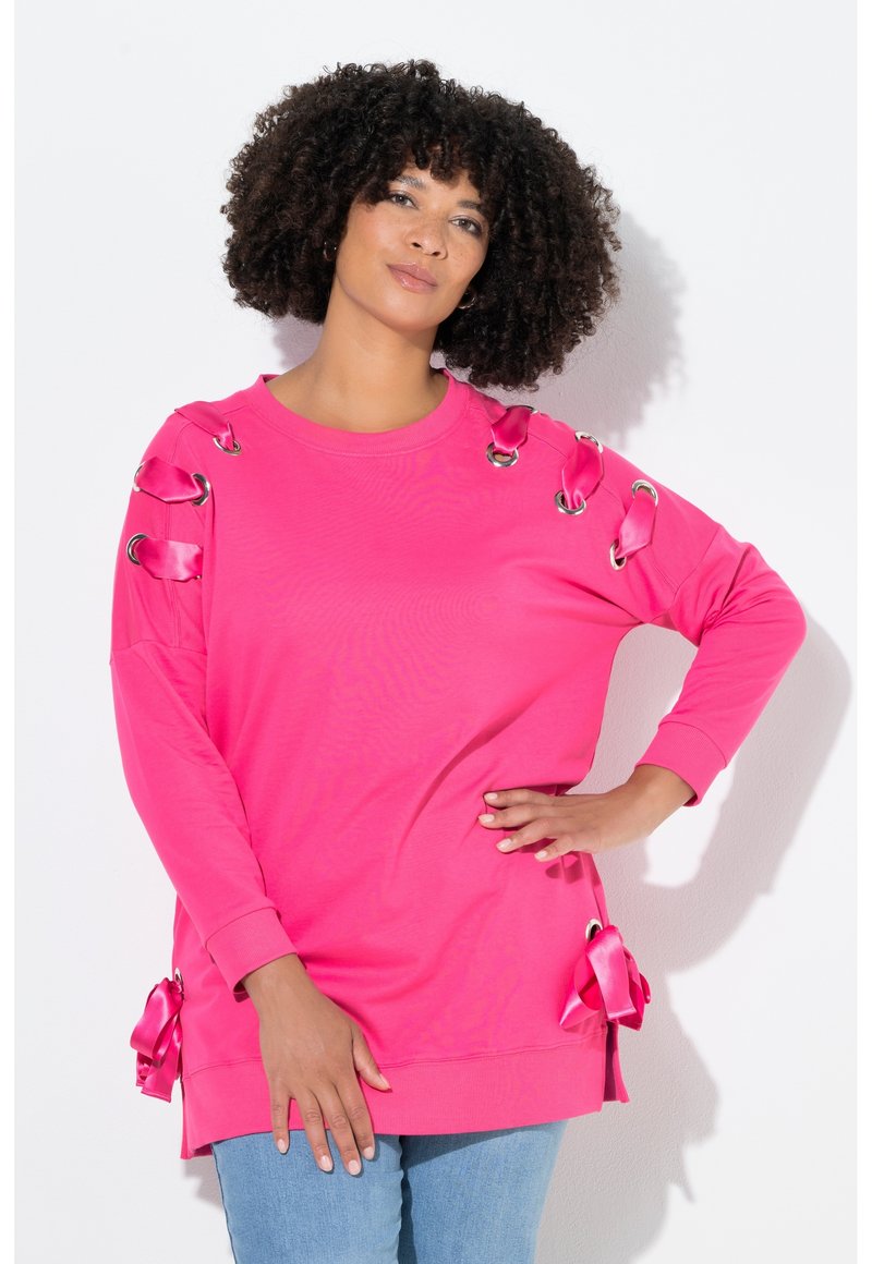 Roze sweater met lange mouwen gemaakt van zachte stof, met decoratieve oogjes en satijnen lintaccenten op de schouders en zijkanten.