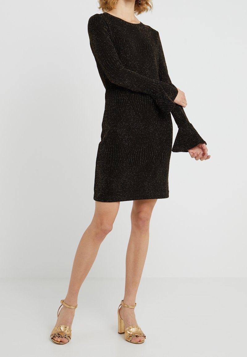 Femme portant une robe noire à manches longues, longueur genou, avec un léger éclat et des sandales dorées à talons hauts, se tenant devant un fond uni.