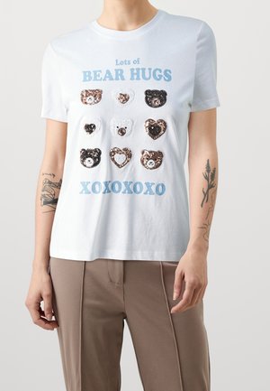 Osoba nosząca białą koszulkę z niebieskim napisem "Lots of BEAR HUGS XOXOXOXO" i dziewięcioma cekinowymi kształtami niedźwiadków i serc z przodu.