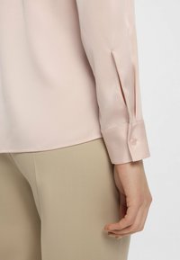 Blusa in satin rosa chiaro con una vestibilità aderente, polsino con chiusura a bottone e una delicata apertura laterale. Abbinata a pantaloni in tessuto beige.