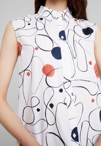 Camicetta bianca senza maniche con linee nere astratte e motivi circolari rossi e blu, completa di chiusura frontale a bottoni e colletto.