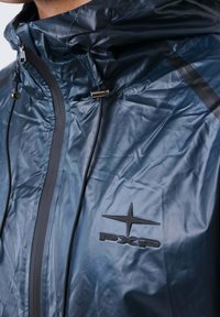 Primo piano di una persona che indossa una giacca impermeabile blu con cappuccio, una zip nera e un logo PXP nero sul petto.