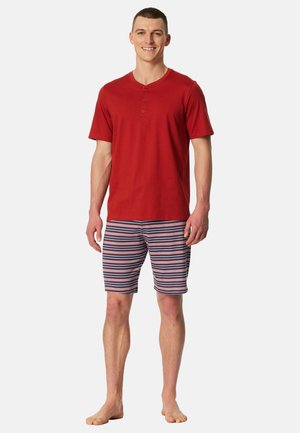 Czerwona koszulka henley z krótkim rękawem i trzema guzikami, w zestawie z paskowanymi szortami w niebieskie, czerwone i białe pasy. Boso, stoi na białym tle.