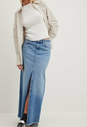 Kvinde iført en ribbet hvid top, beige striped skjorte, lang blå denim nederdel med frontslids, sorte sokker og sneakers, stående mod en ensfarvet baggrund.