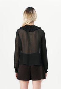 Kvinne med langt blondt hår har på seg en gjennomsiktig svart bluse med lange ermer og volantkrage, samt mørkebrun shorts, vendt bort fra kameraet på hvit bakgrunn.