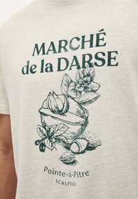 Lichtgrijze t-shirt met een donkergroene illustratie van een kom met noten en bladeren. De tekst luidt "MARCHÉ de la DARSE Pointe-à-Pitre SCALPERS."