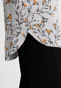 Blusa blanca con patrón floral gris y detalles en naranja. El dobladillo es curvado, mostrando una tela negra debajo. Textura suave.