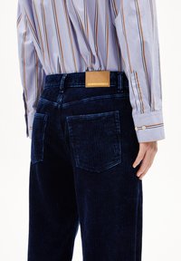 Mörkblå kordflöjelbyxor med vertikala ribs, två bakfickor och en beige läderetikett med varumärket. Randig skjorta synlig ovanför.