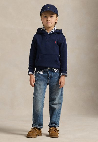 Jeune garçon portant un sweat à capuche bleu marine, une chemise rayée, un jean bleu, des chaussures marron et une casquette bleu marine, debout sur un fond neutre.