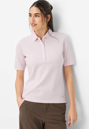 Frau mit dunklem, zu einem Pferdeschwanz gebundenem Haar trägt ein hellrosa kurzärmeliges Poloshirt und braune Hose, steht mit den Händen in den Taschen und schaut nach links.