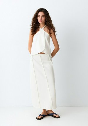 Mujer con cabello rizado, vestida con un top halter blanco y una falda larga, de pie descalza con sandalias negras sobre un fondo de piso y pared blancos.