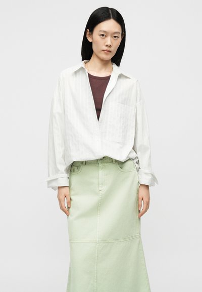 Jeune femme aux cheveux noirs lisses porte une chemise blanche oversize sur un haut marron et une longue jupe vert clair, debout devant un fond gris.