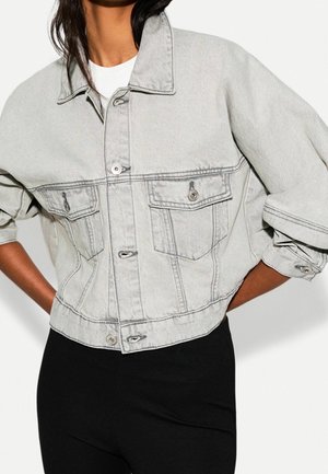 Femme portant une veste en jean gris clair raccourcie avec des poches à boutons et des manches retroussées, associée à un pantalon noir.