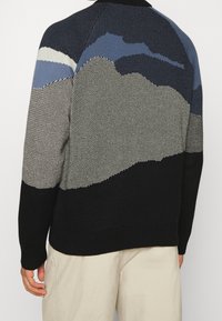 Homme portant un pull en tricot avec un motif abstrait de montagnes en noir, bleu, gris et blanc, assorti à un pantalon beige clair.