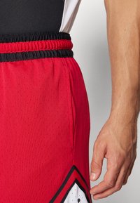 Czerwone spodenki sportowe z czarnym elastycznym pasem, tekstura siateczkowa, boczne kieszenie oraz biały i czarny geometryczny wzór akcentowy.