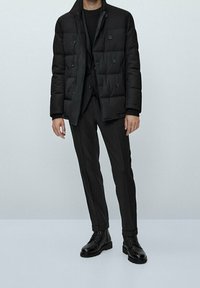 Giacca nera imbottita con colletto alto e doppio bottone, abbinata a pantaloni neri slim-fit e scarpe in pelle nere. Tessuto testurizzato.