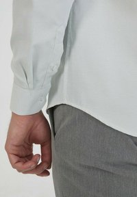 Chemise de ville en gris clair texturé avec des poignets de manche à boutons, associée à un pantalon gris. Main visible reposant près de l'ourlet de la chemise.