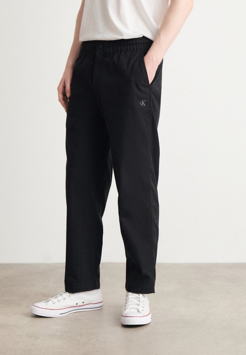 Personne portant un pantalon décontracté noir avec le logo Calvin Klein sur la cuisse, des baskets blanches et une chemise blanche, debout sur un sol uni.