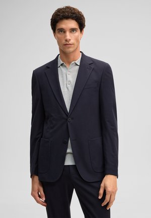 Młody mężczyzna z kręconymi włosami, ubrany w ciemnogranatową marynarkę na jasnoszarej, zapinanej na guziki koszulce polo, stojący na tle jednolitego jasnego tła.