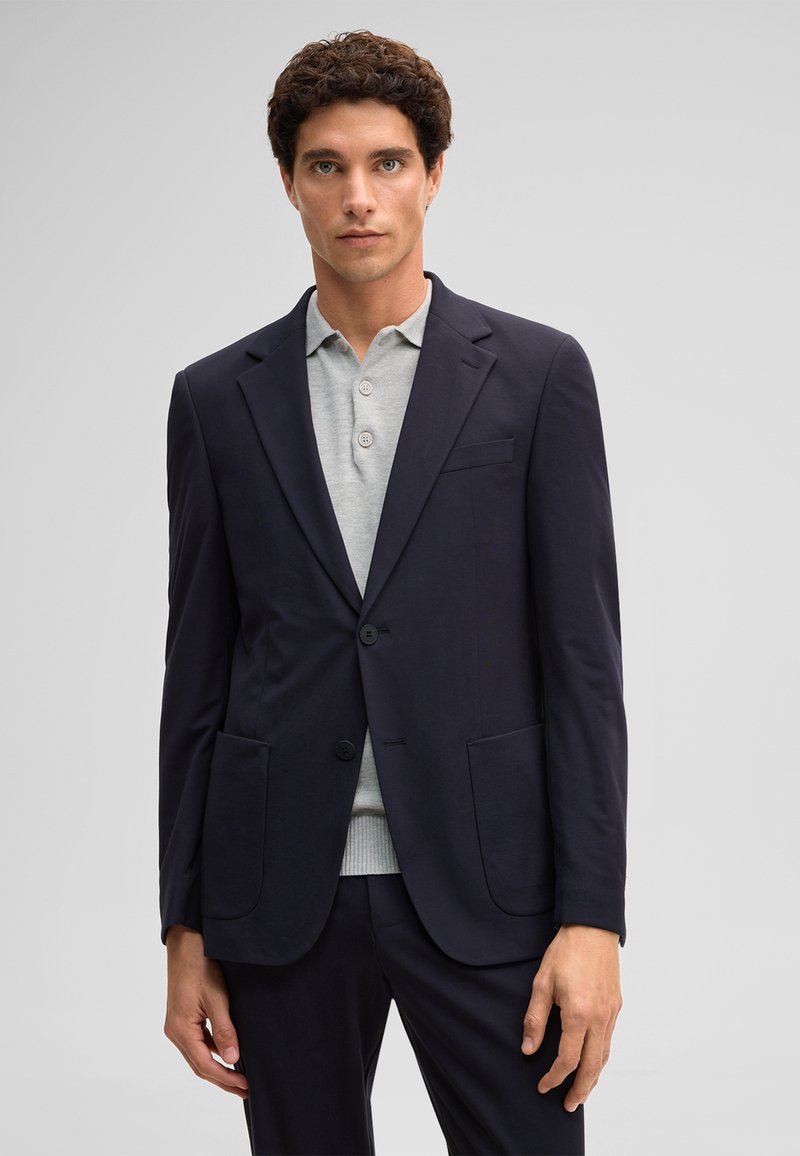 Giovane uomo con capelli ricci che indossa un blazer blu scuro sopra una maglia polo grigio chiaro abbottonata, in piedi davanti a uno sfondo chiaro e uniforme.