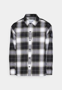 Camicia in flanella a quadri bianco e nero con chiusura a bottoni, due tasche sul petto e un piccolo logo sulla manica sinistra. Texture morbida.