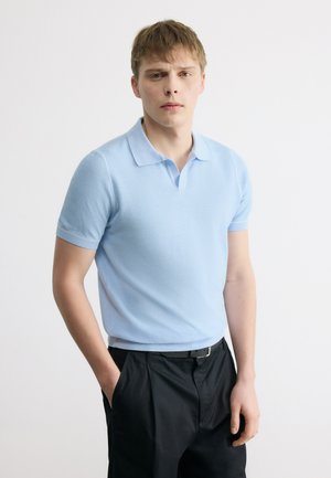 Giovane uomo che indossa una polo a maniche corte azzurro chiaro e pantaloni neri, in piedi con una mano in tasca davanti a uno sfondo bianco semplice.