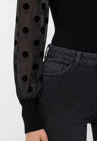 Top nero a coste con maniche a sbuffo trasparenti con pois in velluto nero, abbinato a jeans di denim scuri. Vestibilità slim con tasche.