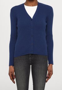 Knapp-up marinblå cardigan med ribbad textur, v-ringad design, långa ärmar och sex knappar fram, kombinerad med mörka jeans.